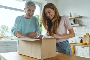Two women stand at a kitchen counter, smiling as they open a Wheaton cancer care package together.