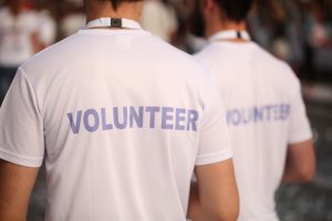 Two people wearing white t-shirts with "VOLUNTEER" printed on the back stand outdoors in a crowd, highlighting the spirit of Wheaton volunteer opportunities.