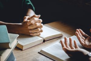Two people sit at a table with open books, one’s hands clasped and the other gesturing, suggesting a discussion about Wheaton cancer care packages or ways to organize Indianapolis donations.