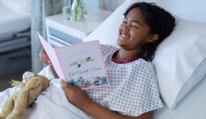 A young girl in a hospital bed smiles while reading a "Get Well Soon" card, holding a stuffed animal. She enjoys the comfort of Wheaton cancer care packages during her stay.
