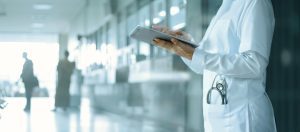 A medical professional in a white coat uses a tablet in a hospital corridor, with a stethoscope in their pocket and people visible in the background—dedicated to delivering Wheaton cancer care packages to those in need.