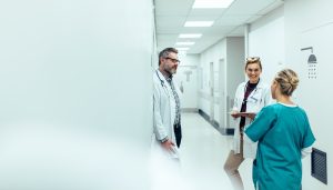Three medical professionals, two in white coats and one in scrubs, stand in a hospital hallway discussing upcoming Crown Point volunteer opportunities.