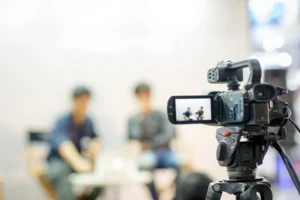 A video camera on a tripod records two people sitting and talking in a blurred background setting.