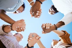 A group of people stand in a circle holding hands, photographed from below against a clear blue sky—united in support, much like those assembling Wheaton cancer care packages for families in need.