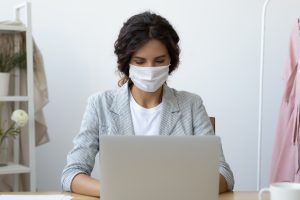A woman wearing a mask sits at a desk and works on a laptop in a bright, indoor setting, researching Wheaton cancer care packages and Wheaton volunteer opportunities to support her community.