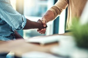 Two people shaking hands across a table in an indoor setting, with a blurred background suggesting an office or meeting space—perhaps connecting over Wheaton volunteer opportunities.
