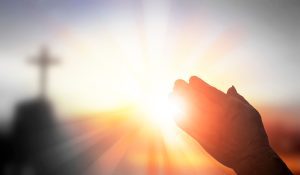 A hand held up toward sunlight with a blurred cross in the background, suggesting a moment of prayer or reflection, evokes the spirit behind Crown Point volunteer opportunities.