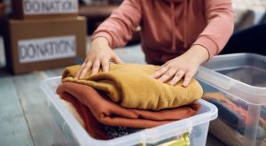 A person folds sweaters into a clear storage bin, preparing Indianapolis donations. Boxes labeled "donation" are visible in the background.