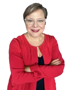 A woman with short brown hair and glasses, wearing a red jacket over a black top, stands with her arms crossed and smiles at the camera—an advocate for Chicago faith-based cancer support.