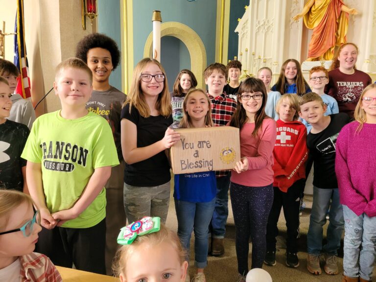 A group of smiling children and teens stand indoors. Two girls at the center hold a cardboard sign that reads, "You are a Blessing," as they gather items for Wheaton cancer care packages.