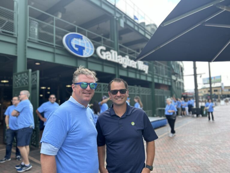 Two men wearing sunglasses and blue shirts stand in front of Gallagher Way, with a group of people and a building in the background, possibly discussing Wheaton volunteer opportunities.