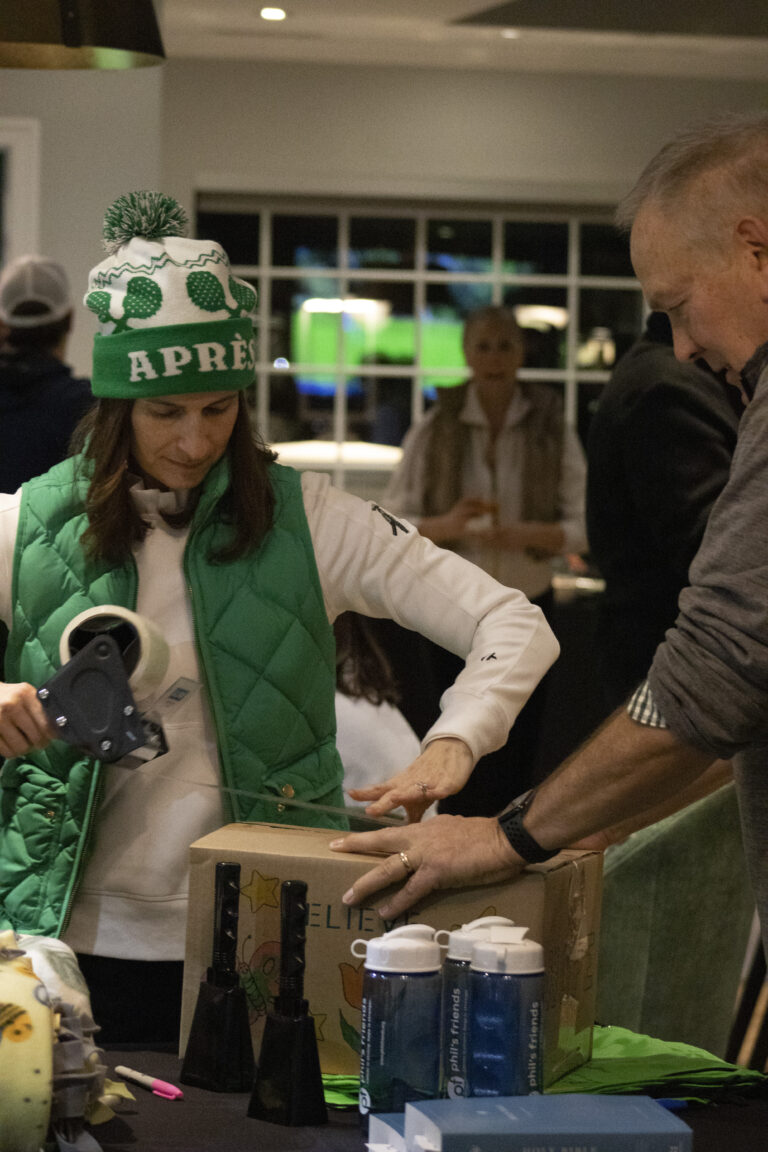 A woman in a green winter hat and vest tapes a cardboard box while a man steadies it; water bottles and other items are on the table during Wheaton cancer care package assembly.
