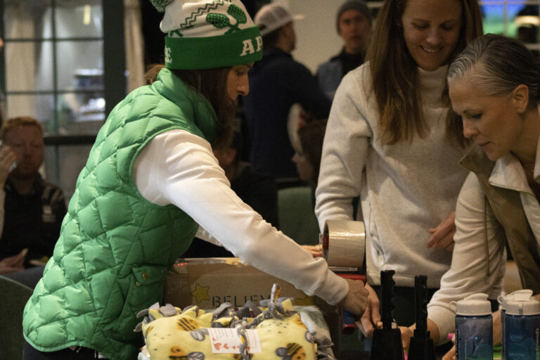 A woman in a green vest and beanie seals a box with tape at a table covered with blankets and bottles, preparing Orlando care packages, while another woman assists. Several people are seated in the background.