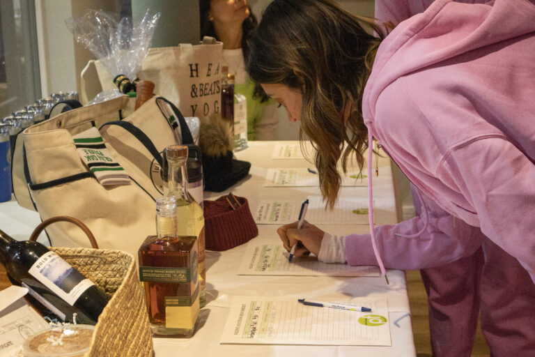A woman in a pink outfit leans over a table to write on a sheet of paper, surrounded by bottles, bags, and auction or raffle items at a Chicago faith-based cancer support event.
