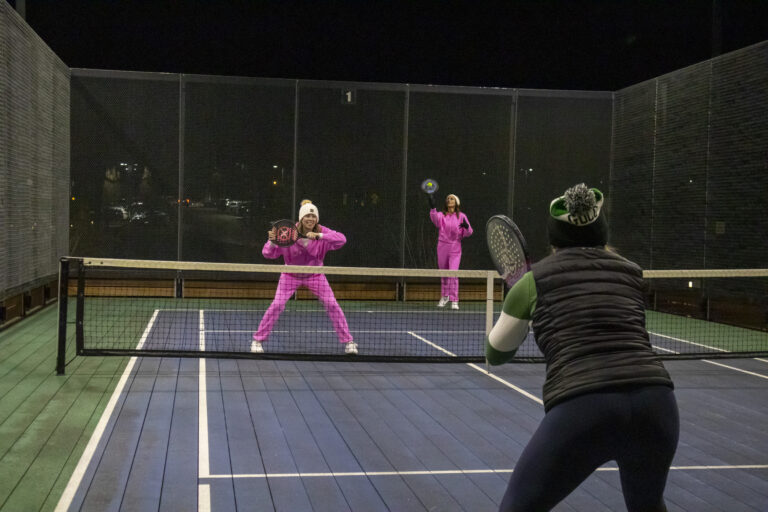 Three women play paddle tennis at night; two wear matching pink outfits, while the third wears a black vest and green hat, showcasing their support for Wheaton cancer care packages.