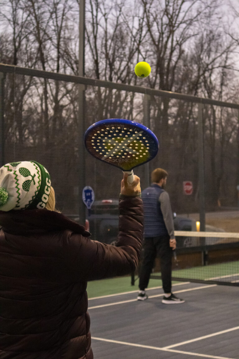 Person in a winter hat prepares to hit a pickleball with a paddle on an outdoor court, while another player stands near the net—just one way to connect, like exploring Crown Point volunteer opportunities in your community.