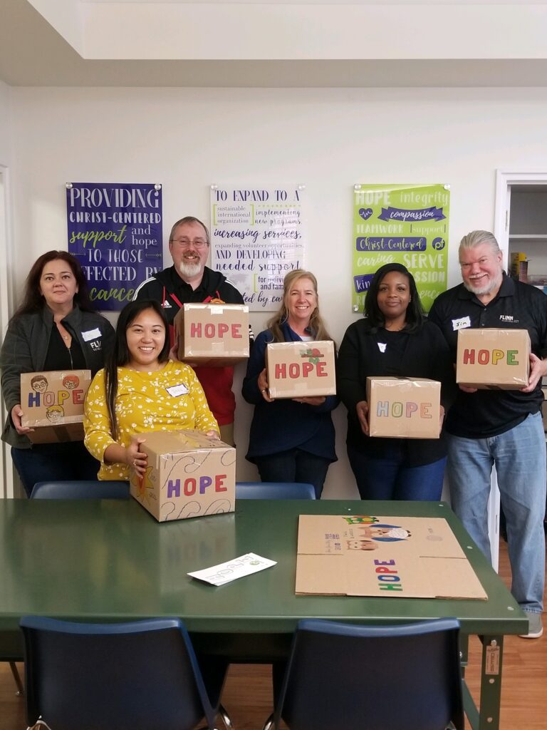 Six people stand indoors holding cardboard boxes labeled "HOPE," showcasing Wheaton cancer care packages, with posters and a green table visible in the background.