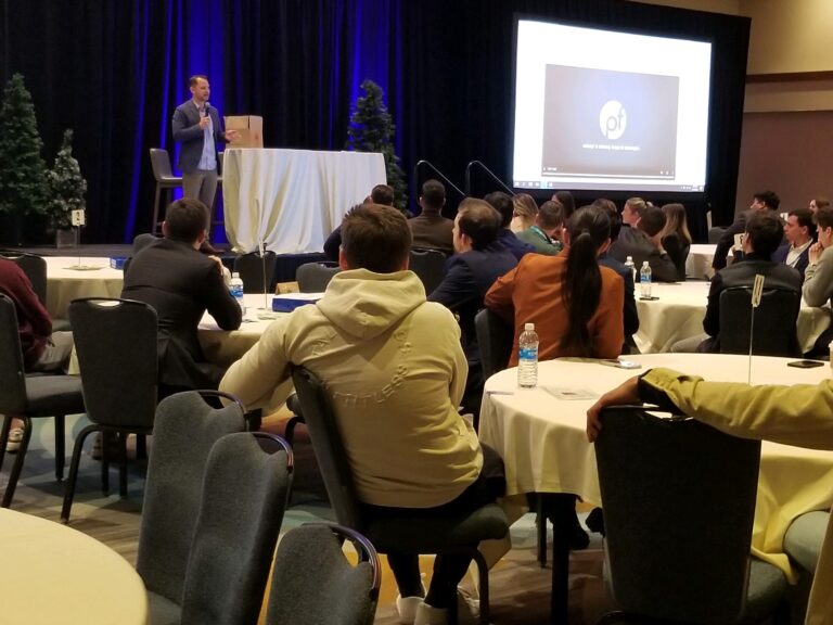 A man in business attire speaks on stage to an audience seated at round tables, sharing insights about Wheaton cancer care packages, with a presentation projected on a screen behind him.