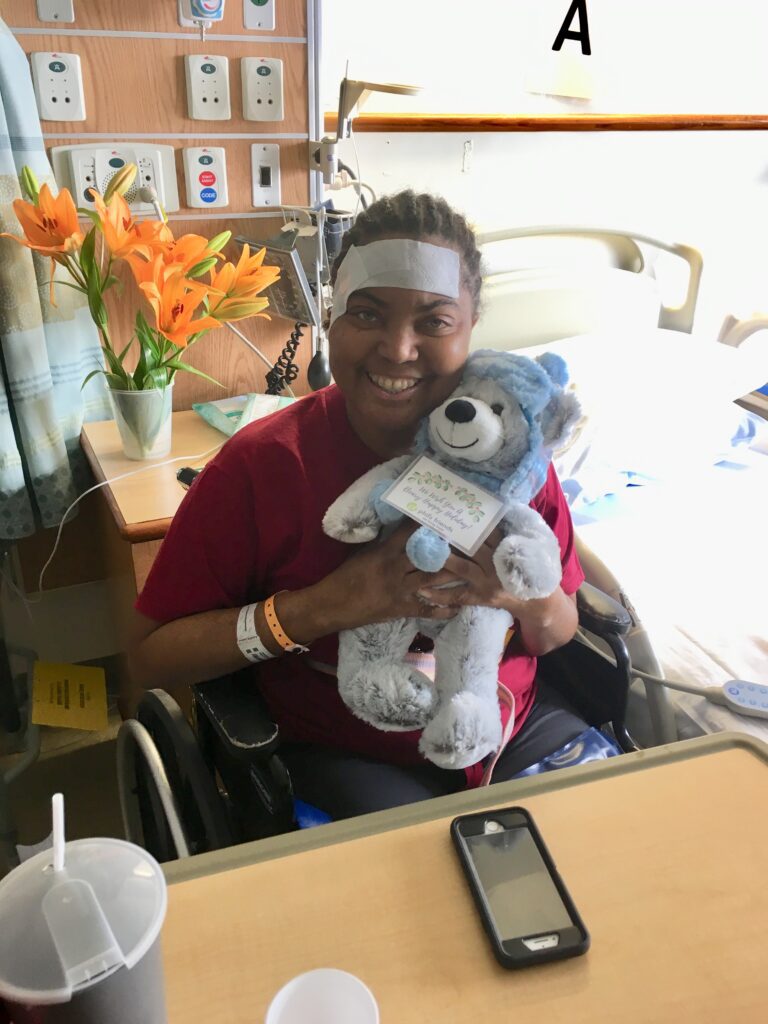 A person in a hospital room sits in a wheelchair, smiling and holding a blue teddy bear. Hospital equipment, flowers, and a bedside table are visible in the background—reminding us of Crown Point volunteer opportunities to lift patients' spirits.