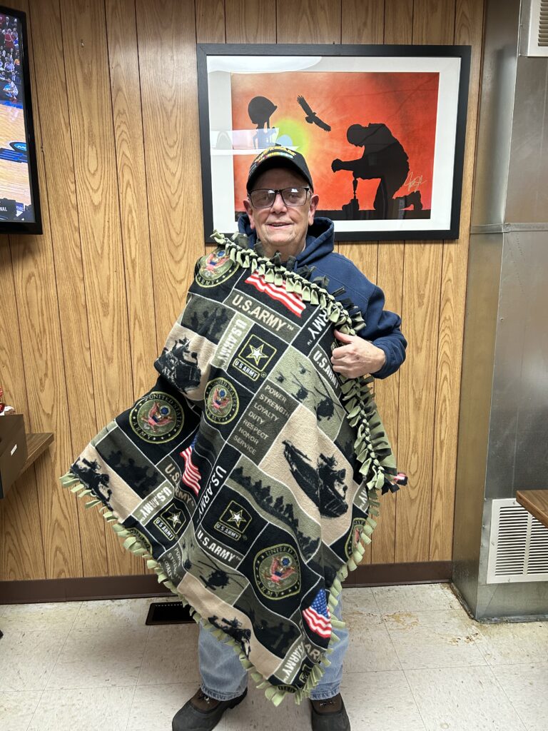 An older man in a cap and glasses stands indoors holding a U.S. Army-themed blanket, part of Wheaton cancer care packages. Wood-paneled walls and framed art are visible behind him.