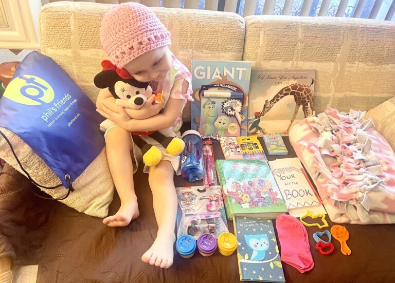 A young child wearing a pink hat sits on a couch hugging a Minnie Mouse toy, surrounded by books, toys, Play-Doh, a fleece blanket, and a blue "Phil's Friends" bag—a symbol of Chicago faith-based cancer support.