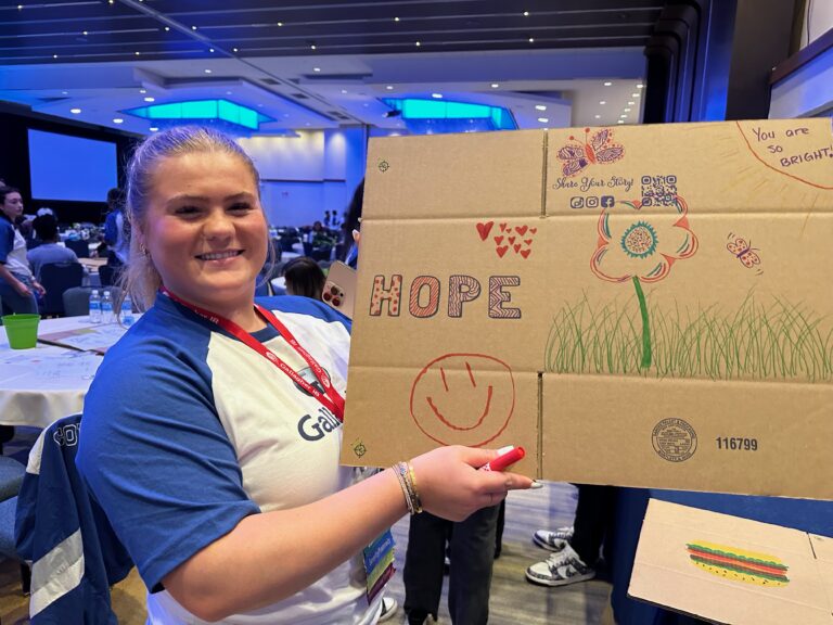 A woman smiles and holds a cardboard sign decorated with drawings, the word "HOPE," a smiley face, and colorful designs in a conference room, spreading cheer while preparing Wheaton cancer care packages.