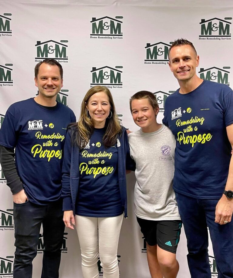 Four people stand and smile in front of an M&M Home Remodeling Services backdrop. Two adults wear matching "Remodeling with a Purpose" shirts, celebrating Wheaton volunteer opportunities.