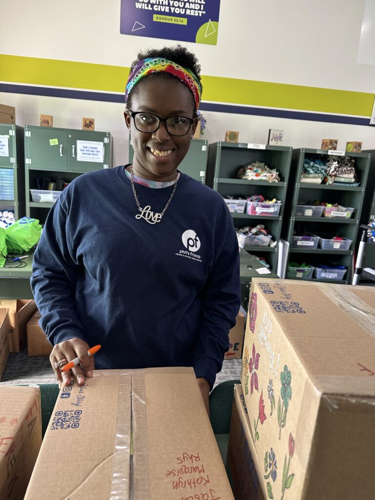 A person wearing a blue sweatshirt and rainbow headband smiles while packing boxes for Wheaton cancer care packages in a room with shelves and supplies.