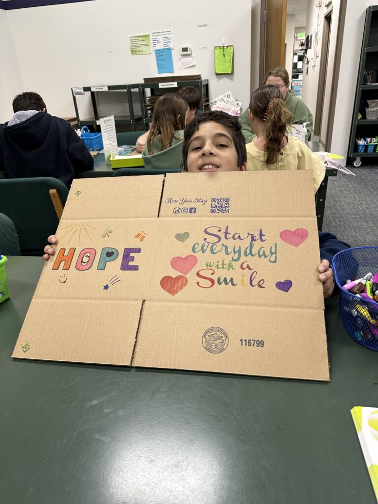A boy holds a cardboard sign decorated with colorful letters, hearts, and the message “HOPE Start everyday with a Smile” in a classroom—showcasing the impact of Wheaton cancer care packages on brightening lives and spreading positivity.