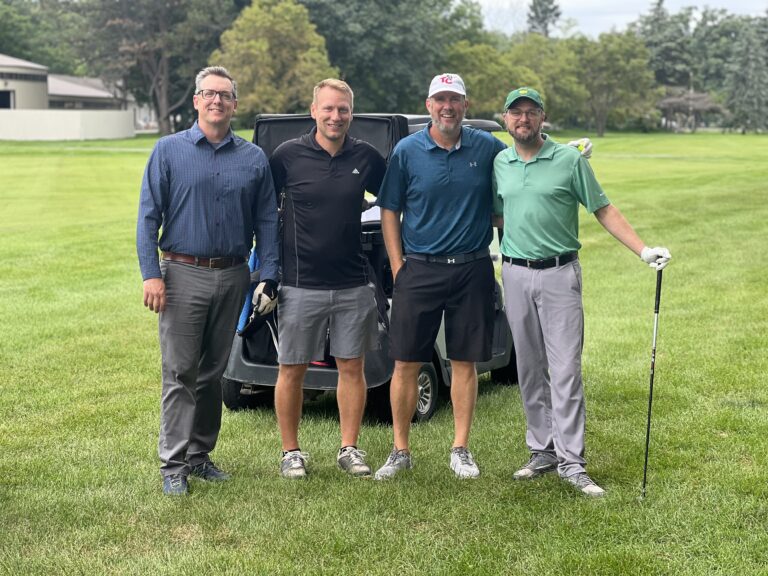 Four men stand side by side on a golf course with a golf cart behind them; three are in golf attire holding clubs, while one, in business casual, supports Chicago faith-based cancer support initiatives.