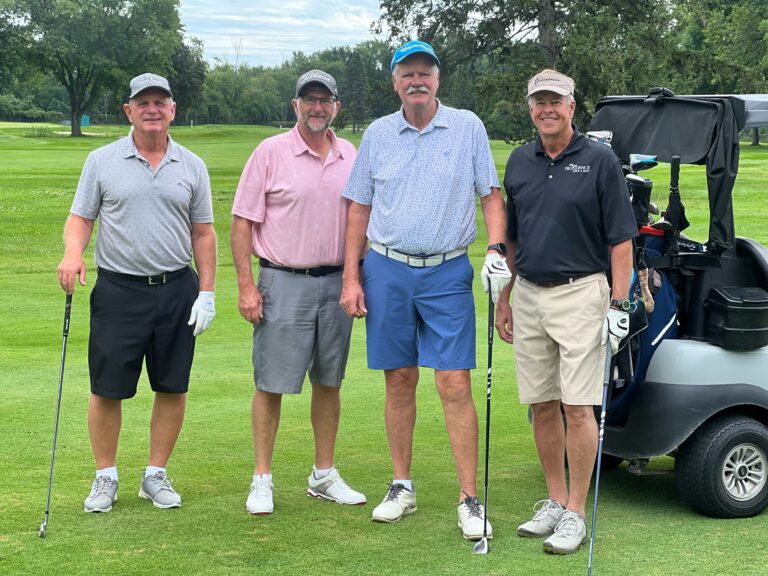Four men stand on a golf course near a golf cart, holding clubs and dressed in casual golf attire. They're enjoying the day while discussing Wheaton volunteer opportunities. Lush trees and grass create a picturesque background.