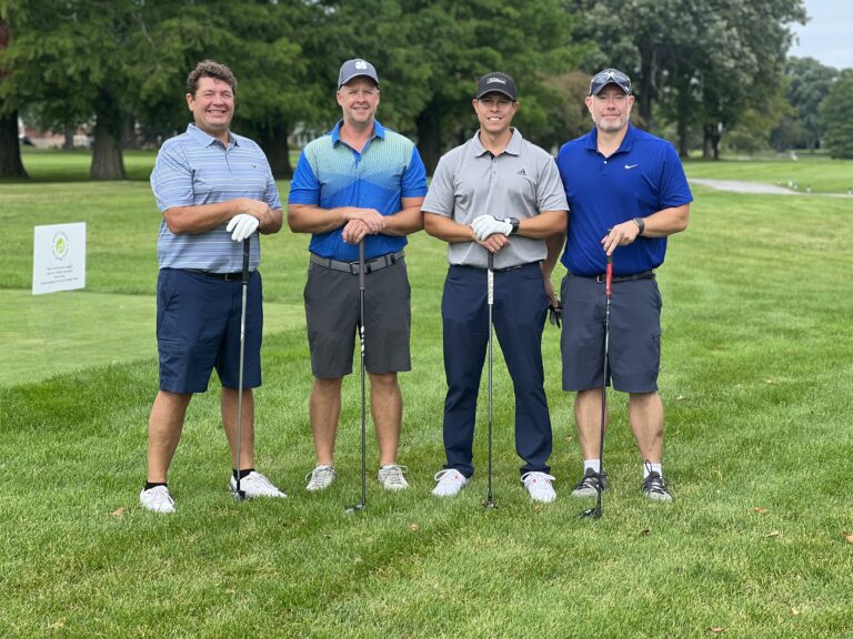 Four men stand on a golf course holding clubs, posing for a group photo on the grass with trees and a golf sign in the background, supporting Chicago faith-based cancer support through their participation.