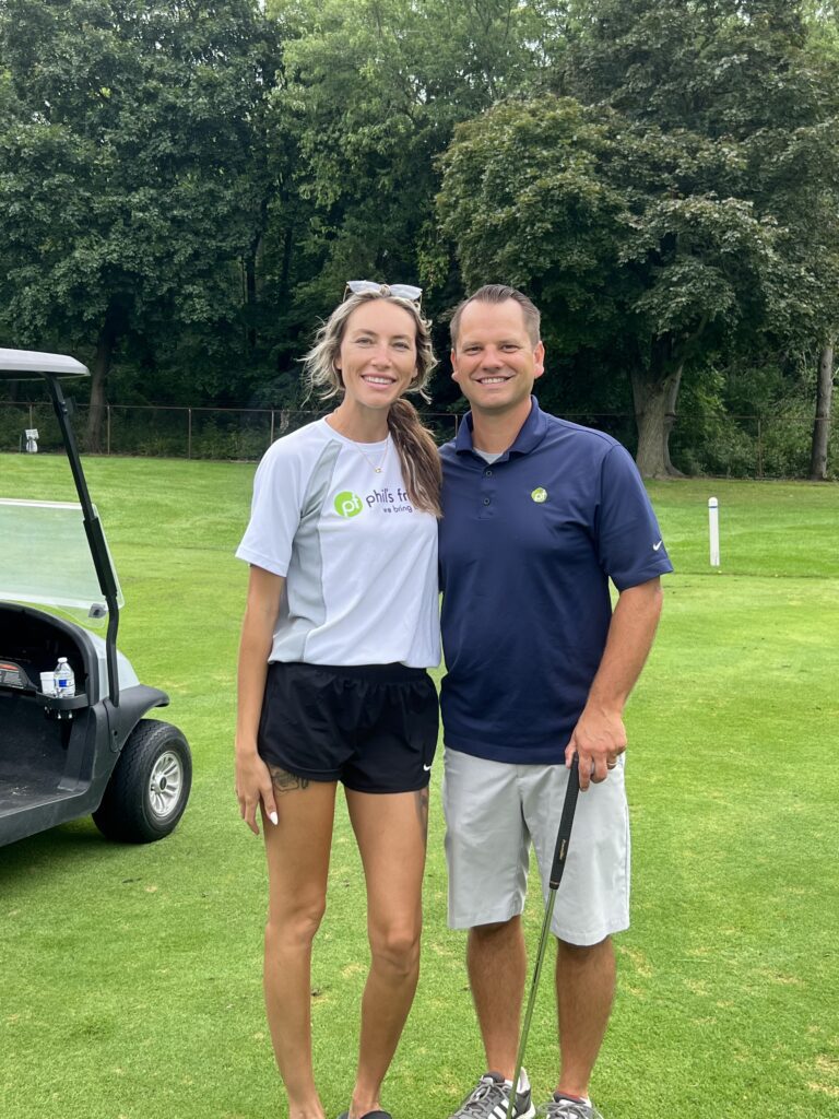 A woman and a man stand together on a golf course near a golf cart, enjoying their day after connecting through Wheaton volunteer opportunities. The man holds a golf club and both are smiling. Trees and grass are visible in the background.