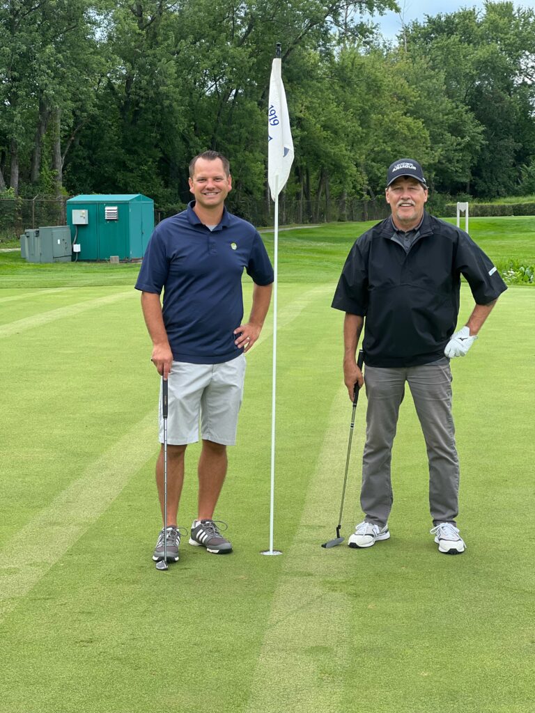 Two men stand on a golf green near the hole flag, each holding a putter. Trees and a portable toilet are visible in the background, possibly at an event supporting Crown Point volunteer opportunities.