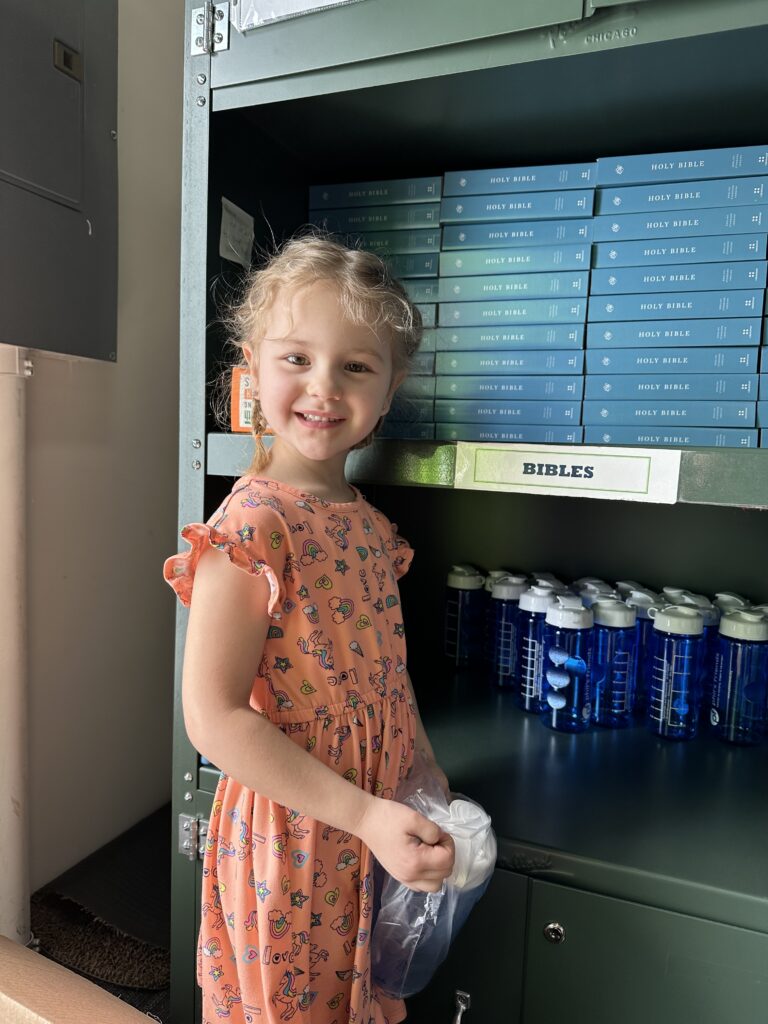 A young girl in an orange patterned dress stands in front of a shelf with stacks of blue books labeled "Bibles" and several blue water bottles, representing Crown Point volunteer opportunities.