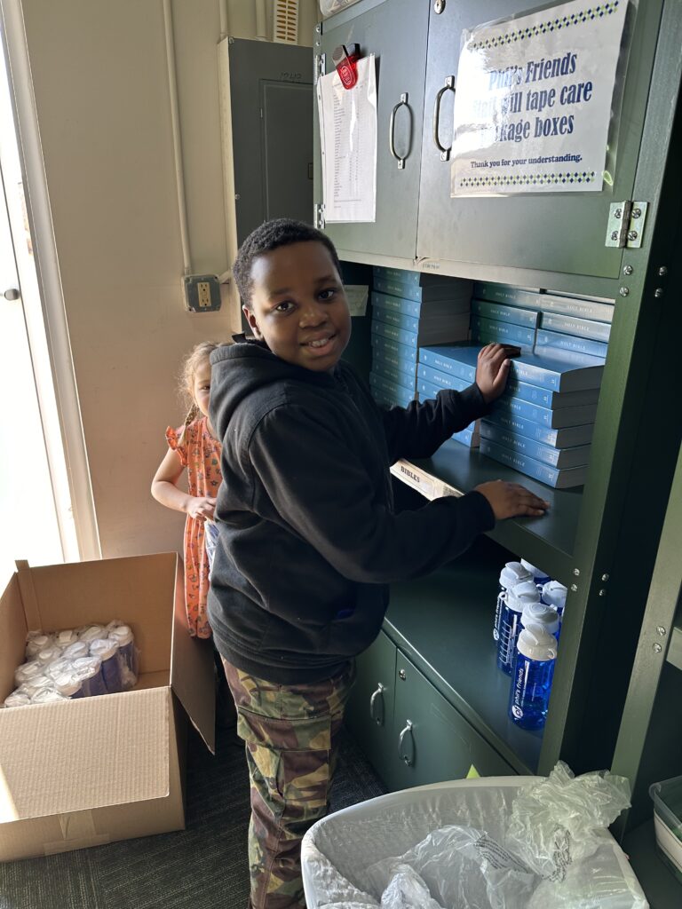 A boy organizes blue books on a shelf while a girl stands nearby, helping with Wheaton cancer care packages. Bottles of water and a box of packaged items are on the floor.