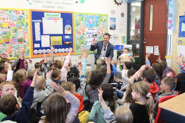 A teacher sits at the front of a classroom, holding a book and speaking to a large group of young students who are seated on the floor and raising their hands—highlighting the spirit found in Wheaton volunteer opportunities.