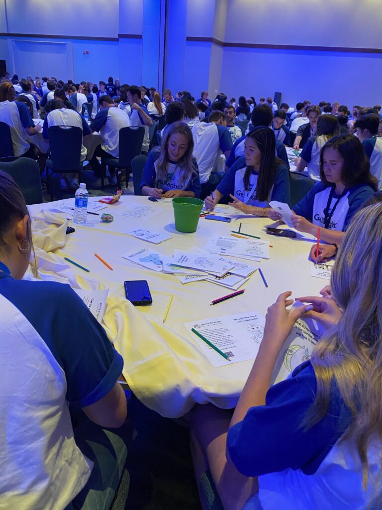 A group of people sit around a table, drawing and writing on papers with colored pencils at an indoor event, assembling Wheaton cancer care packages with many participants.