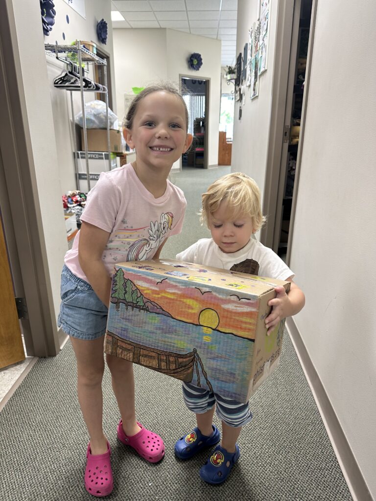 Two children stand in a hallway, holding a cardboard box decorated with a colorful sunset—one of many Wheaton cancer care packages made through local volunteer opportunities.
