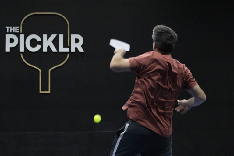 A man in a red shirt hits a pickleball with a paddle on an indoor court, with "The Picklr" logo on the wall, highlighting Indianapolis donations that help support local recreational activities.