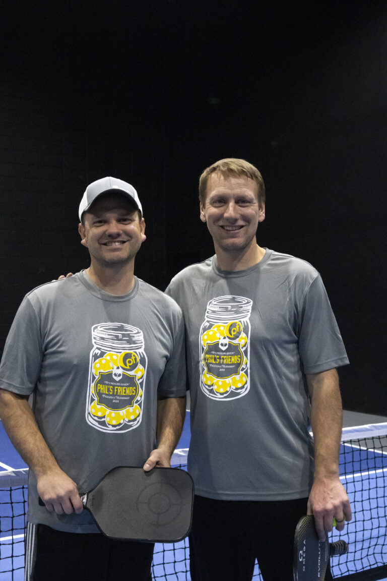 Two men wearing matching gray “Phil’s Friends” pickleball shirts and holding paddles stand side by side on an indoor court, smiling at the camera—highlighting Wheaton volunteer opportunities in support of a great cause.