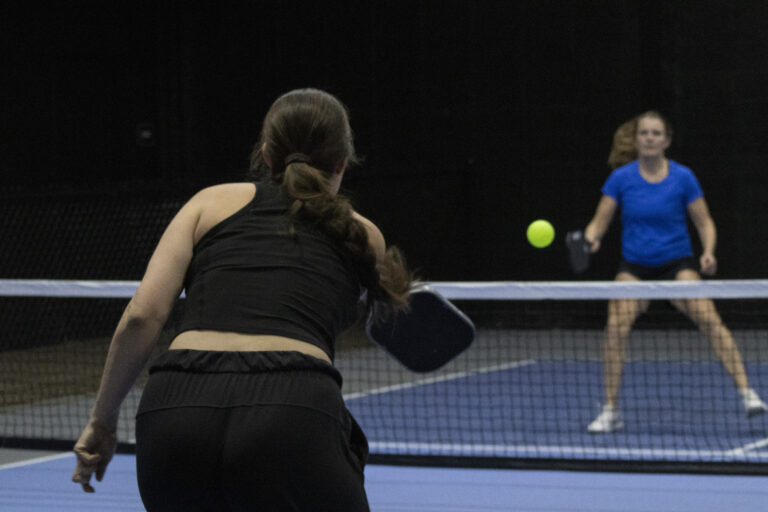 Two women play pickleball on an indoor court, one in black preparing to hit the ball while the other in blue stands ready on the opposite side of the net during a Crown Point volunteer opportunities event.