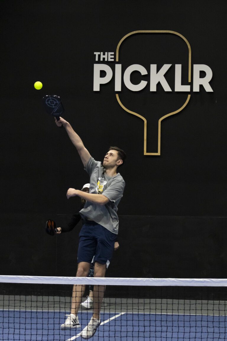 Two people play pickleball indoors; one jumps to hit a ball with a paddle in front of a wall displaying "The Picklr" and a large paddle graphic, supporting Indianapolis donations for local causes.