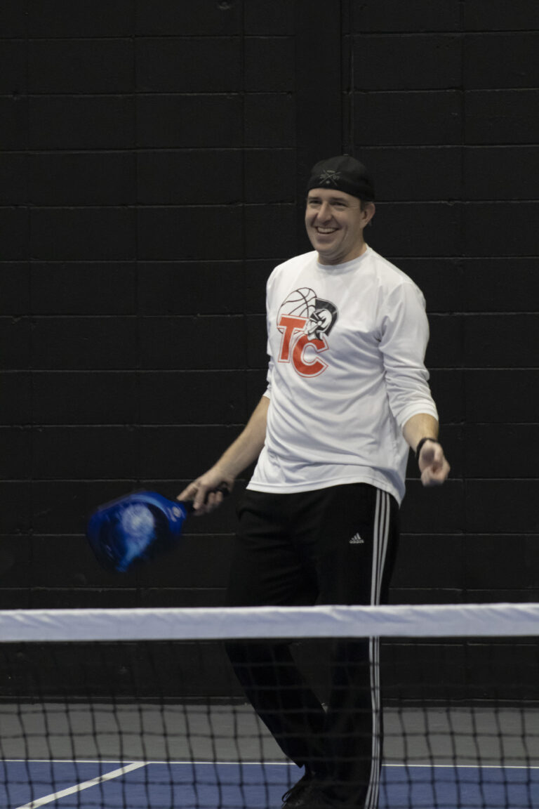 A man wearing a white TC shirt and black pants stands on an indoor court holding a blue pickleball paddle, smiling while raising awareness for Wheaton cancer care packages.