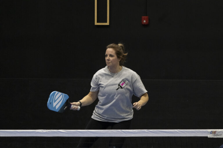 Woman in a gray t-shirt holding a blue pickleball paddle stands on an indoor court, supporting Chicago faith-based cancer support through community events.