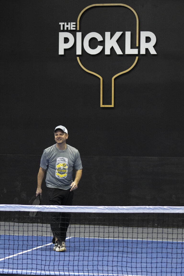 A man holding a pickleball paddle stands on an indoor court in front of a wall with "THE PICKLR" logo and a paddle graphic, highlighting Crown Point volunteer opportunities in the local community.