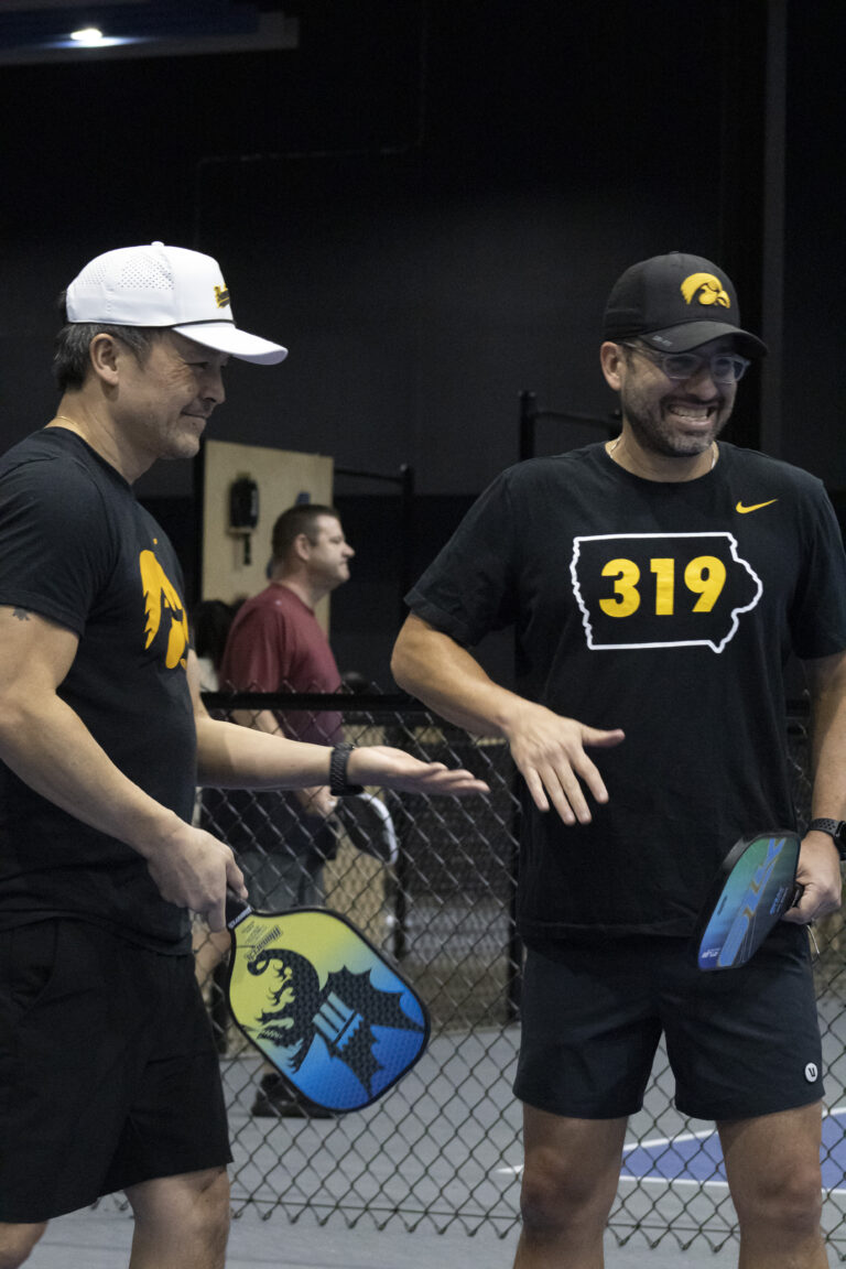 Two men stand on a pickleball court holding paddles, both in black Iowa-themed shirts and caps, supporting Chicago faith-based cancer support. A chain-link fence and another player are visible in the background.