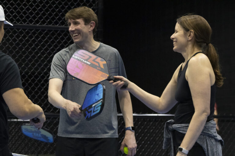 Two people in athletic wear smile and tap pickleball paddles with others on an indoor court, a black chain-link fence behind them—highlighting camaraderie and the spirit found at Crown Point volunteer opportunities.