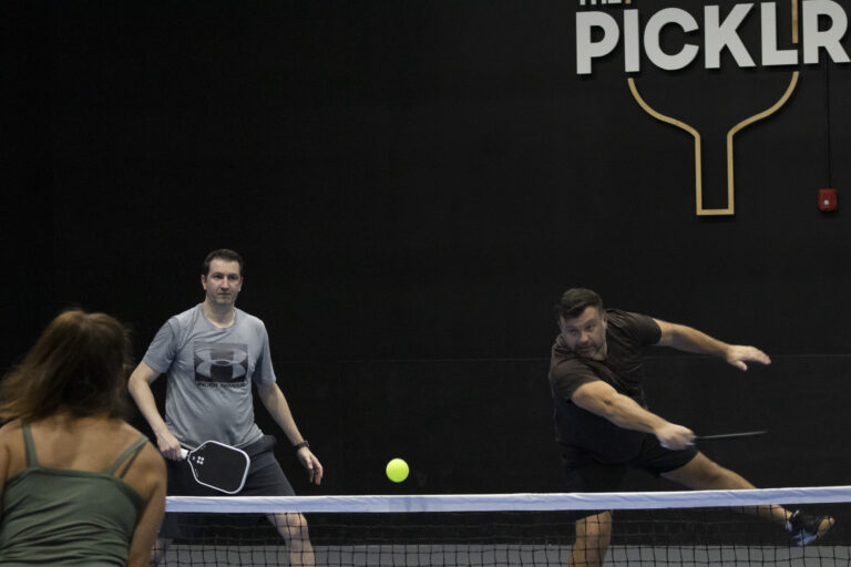 Two men play pickleball indoors; one lunges to hit the ball while the other watches. A woman is partially visible in the foreground. The word "PICKLR" is on the wall, highlighting Crown Point volunteer opportunities in the community.
