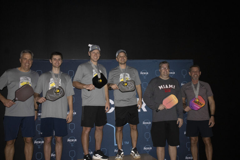 Six men stand in front of a Picklr-branded backdrop holding pickleball paddles, some wearing medals, celebrating teamwork and highlighting the spirit of Crown Point volunteer opportunities.
